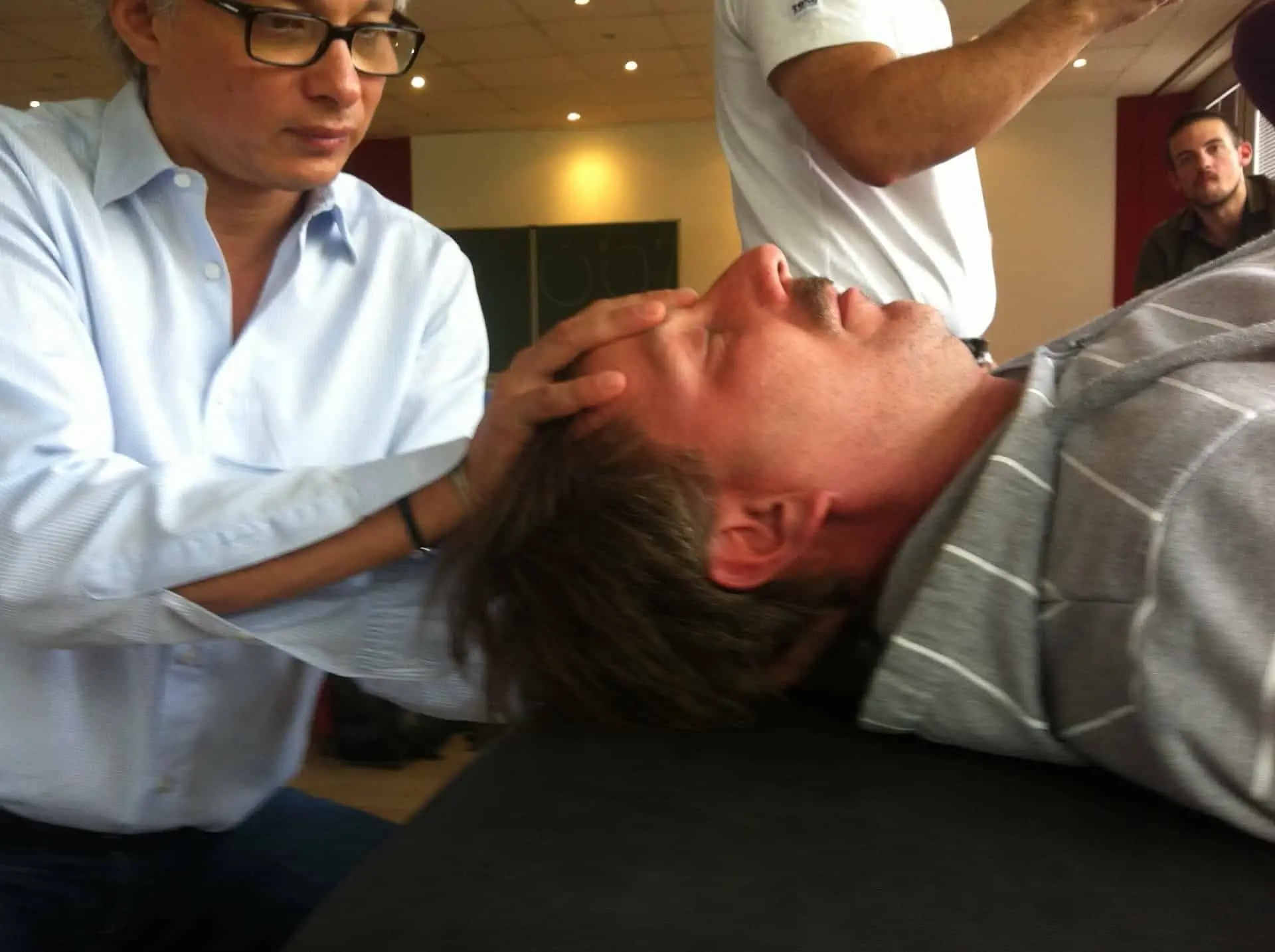 Ein Osteopath in Hamburg behandelt einen Mann osteopathisch, der auf einem Bett liegt und dessen Kopf auf einem Tisch liegt.