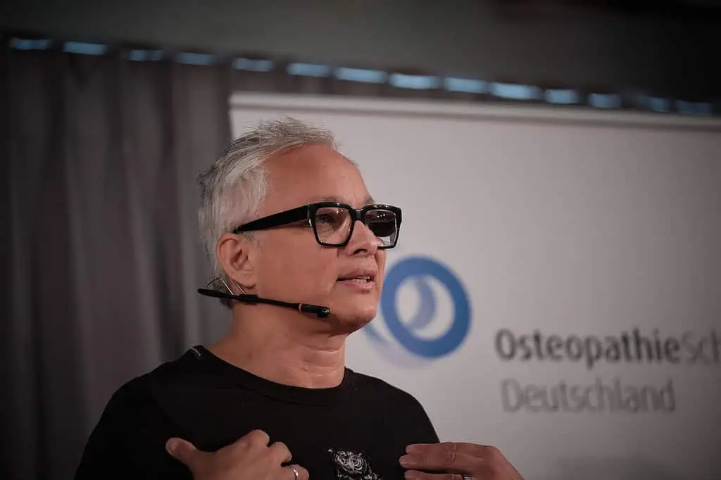 Ein Osteopath mit Brille steht vor einem Schild.