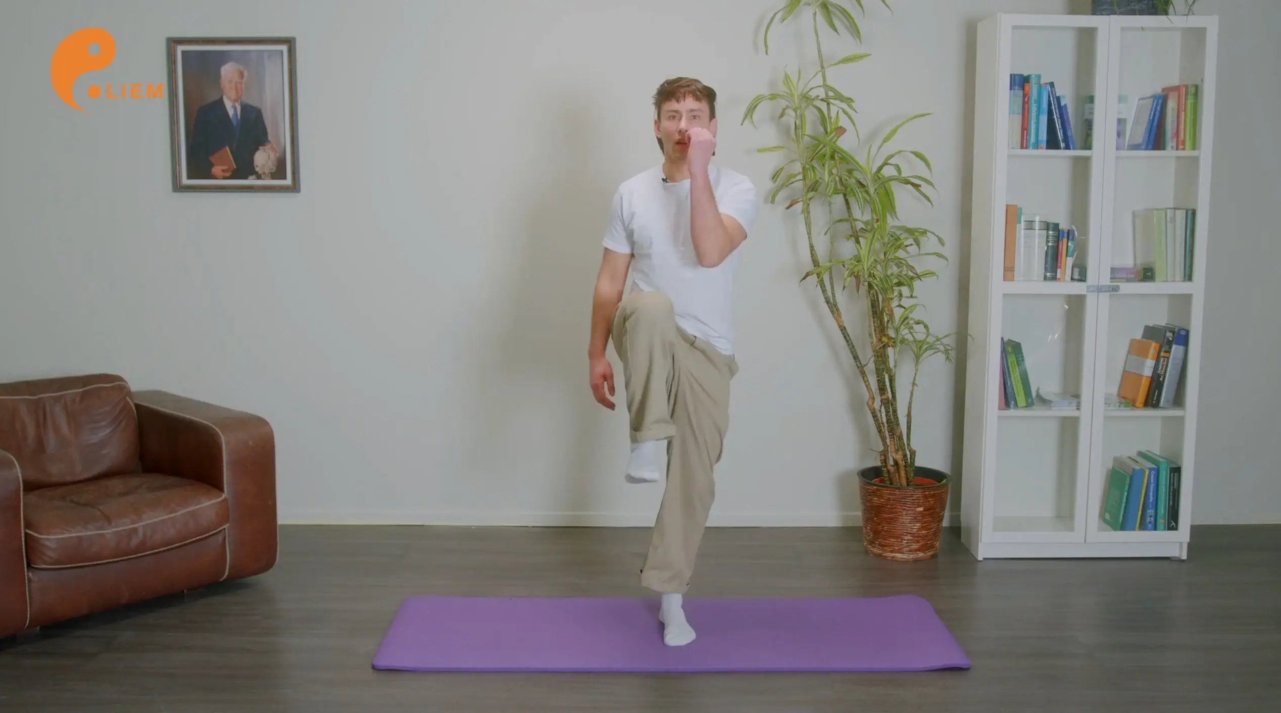 Eine Person macht drinnen auf einer lila Yogamatte Übungen. Sie macht High Knees, indem sie ihr rechtes Knie anhebt und es mit der linken Hand berührt. Sie trägt ein weißes T-Shirt und beige Hosen. Hinter ihr stehen eine Topfpflanze, ein Bücherregal, ein Ledersessel und ein gerahmtes Foto.