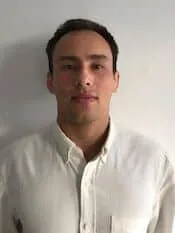 Ein Mann mit kurzen dunklen Haaren steht vor einem schlichten hellen Hintergrund und blickt direkt in die Kamera. Er trägt ein helles Hemd mit Knöpfen und verkörpert damit das professionelle Auftreten, das man von einem Osteopathie-Arzt in Hamburg erwarten würde.