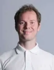 Eine Person mit kurzen, hellbraunen Haaren trägt ein weißes Hemd mit Kragen und lächelt, möglicherweise ein Osteopathie-Experte in Hamburg. Der Hintergrund ist schlicht und hellgrau.