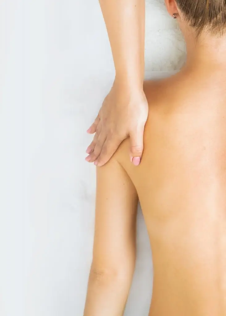 Eine Person mit nacktem Rücken erhält von einem Osteopathen in Hamburg eine Schultermassage. Die mit pinkfarbenen Fingernägeln verzierte Hand auf der Schulter strahlt Sorgfalt und Präzision aus. Der Hintergrund ist eine helle, schlichte Wand, die die beruhigende Atmosphäre der Behandlungssitzung unterstreicht.