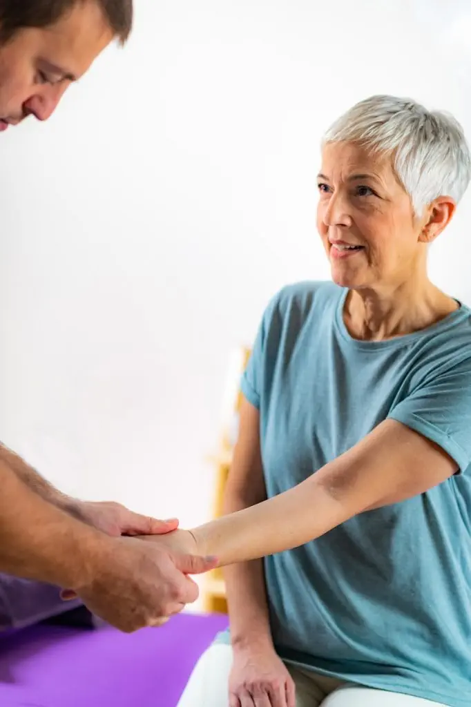 Eine Person mit kurzem grauem Haar und blauem Hemd wird von einer anderen Person mit dem linken Arm gestützt. Die Szene spielt in einem Innenbereich und konzentriert sich auf die Interaktion zwischen den beiden Personen. Dabei wird die mitfühlende Betreuung hervorgehoben, die typisch für die Osteopathie in Hamburg ist.