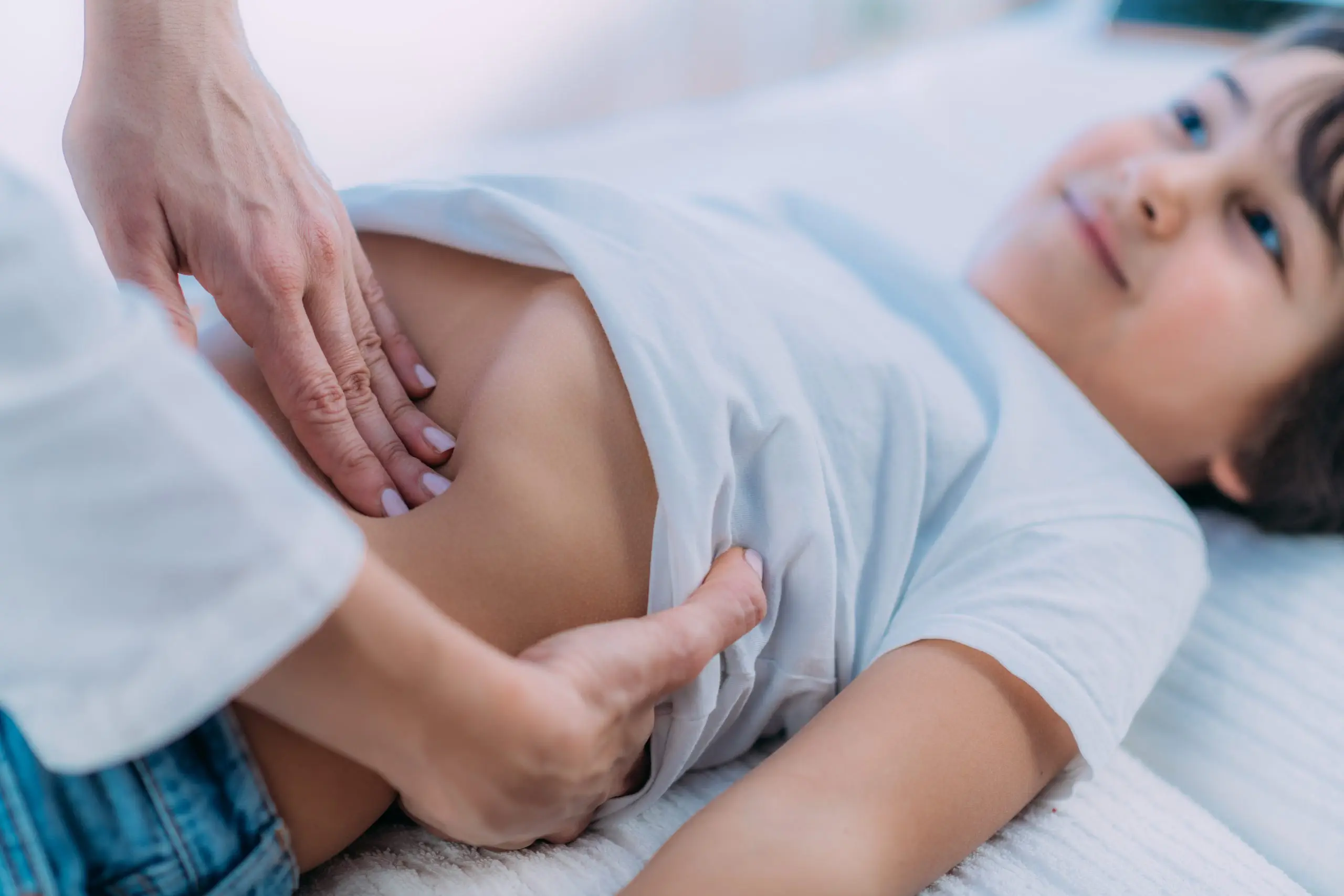 Une personne effectue un examen abdominal sur un enfant allongé sur un lit. L'enfant, vêtu d'une chemise blanche et d'un jean bleu, est souriant et détendu. En plus d'appuyer doucement sur le ventre de l'enfant, l'adulte intègre subtilement des éléments de la technique de la pompe à pédale pour assurer son confort.
