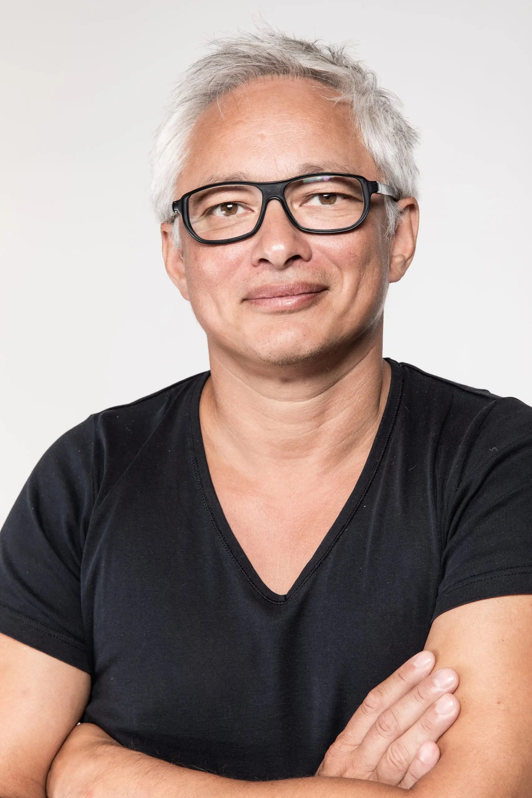 Eine Person mit kurzen grauen Haaren und Brille trägt ein schwarzes V-Ausschnitt-Shirt. Sie blickt mit einem leichten Lächeln in die Kamera und hat die Arme verschränkt. Der Hintergrund ist schlicht weiß.