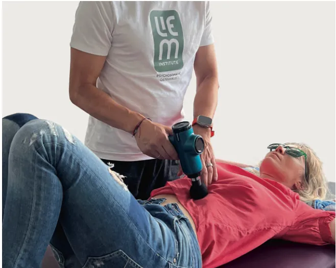 Eine Person in einem weißen Hemd verwendet ein Handmassagegerät für die Sonnengeflechtbehandlung auf dem Bauch einer auf einem Tisch liegenden Frau. Die Frau, die eine Sonnenbrille, ein rotes Hemd und eine blaue Jeans trägt, wirkt während der Behandlung entspannt.